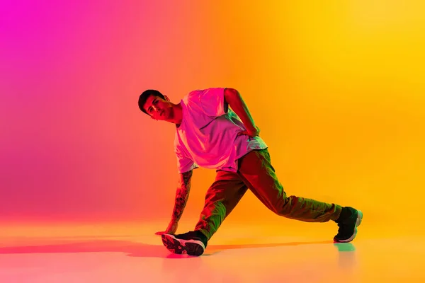 Solo optreden. Jongeman, breakdancing danser tactiek in casual kleding geïsoleerd over verloop roze gele achtergrond in danszaal in neon licht. — Stockfoto