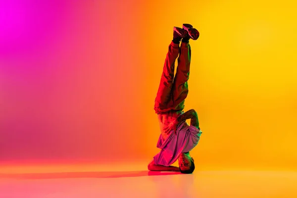 Full length portret van jonge stijlvolle man, break dancing danser in casual kleding geïsoleerd over verloop roze gele achtergrond in danszaal in neon licht. — Stockfoto