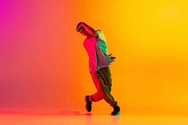 Full length portret van jonge stijlvolle man, break dancing danser in casual kleding geïsoleerd over verloop roze gele achtergrond in danszaal in neon licht. — Stockfoto