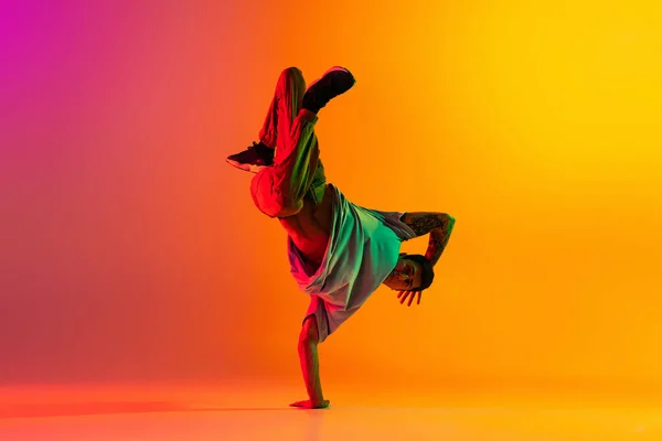 Portret młodego stylowego mężczyzny, break dancing tancerka trening w ubraniach casual odizolowanych nad gradientowym różowym tle żółty w sali tanecznej w neonowym świetle. — Zdjęcie stockowe