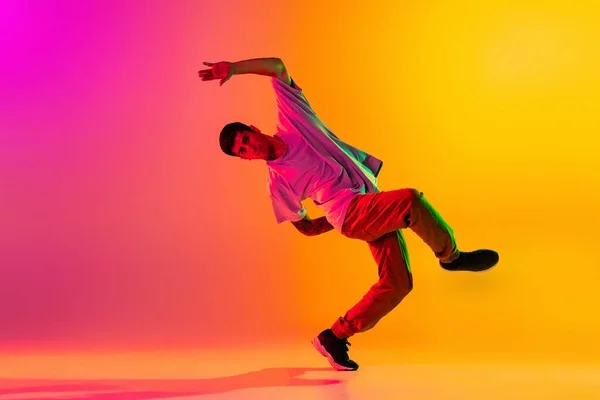 Portret van jonge stijlvolle man, breakdance danseres training in casual kleding geïsoleerd over hellend roze gele achtergrond in danszaal in neon licht. — Stockfoto