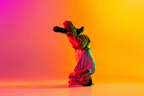 In actie. Jongeman, breakdancing danser tactiek in casual kleding geïsoleerd over verloop roze gele achtergrond in danszaal in neon licht. — Stockfoto