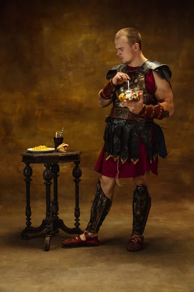 Portrait of medieval person, young man, warrior or knight in war equipment eating isolated on vintage dark background. Comparison of eras, art, history, humor, retro style concept