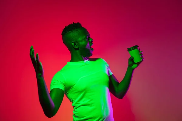 Kreatywny wizerunek afrykańskiego młodzieńca w luźnych ubraniach odizolowanych na czerwonym tle studia w neonowym świetle, filtr. Pojęcie ludzkich emocji, ekspresji twarzy, młodości, uczuć, reklam. — Zdjęcie stockowe