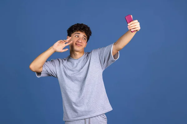 Telefon kullanan genç kıvırcık adamın portresi mavi stüdyo arka planında izole edilmiş ve reklam için kopyalanmış. İnsan duyguları, moda, güzellik, gençlik kavramı — Stok fotoğraf