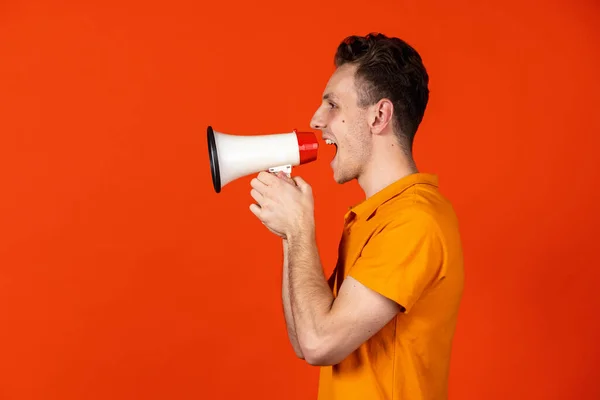 Portrait of young Caucasian man shouting at megaphone isolated on orange color studio background. Concept of human emotion, facial expressions, youth, feelings, ad. — Stock Photo, Image