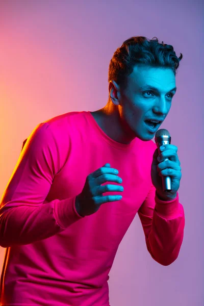 Pół długości portret młodego przystojnego mężczyzny z mikrofonem odizolowany fioletowy kolor studio tło w różowym neonowym filtrze światła. Pojęcie ludzkich emocji, wyrazu twarzy, młodości, uczuć, reklam. — Zdjęcie stockowe