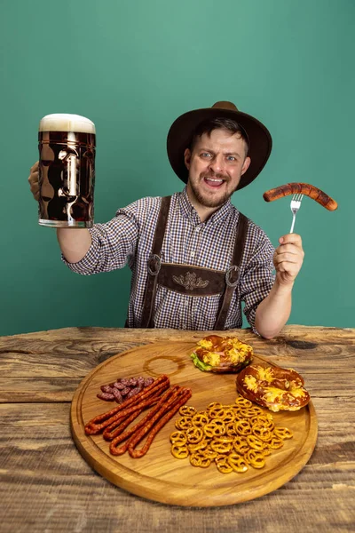 Young man dressed in traditional Austrian or Bavarian costume sitting at table with festive food and beer isolated over green background — Stock Photo, Image