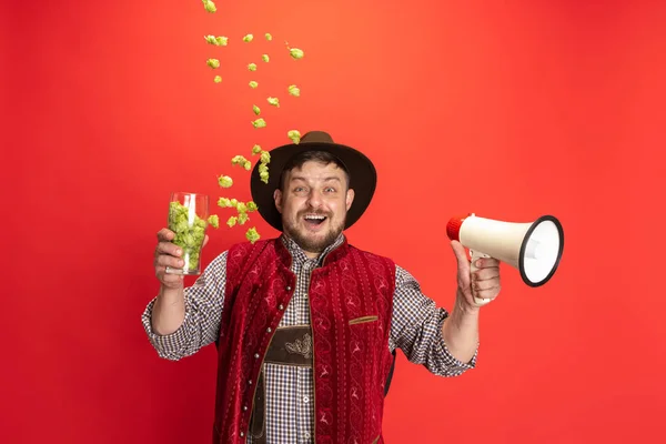Szczęśliwy uśmiechnięty mężczyzna ubrany w tradycyjny austriacki lub bawarski kostium z megafonem i szklankami wypełnionymi dzikim chmielem na czerwonym tle — Zdjęcie stockowe