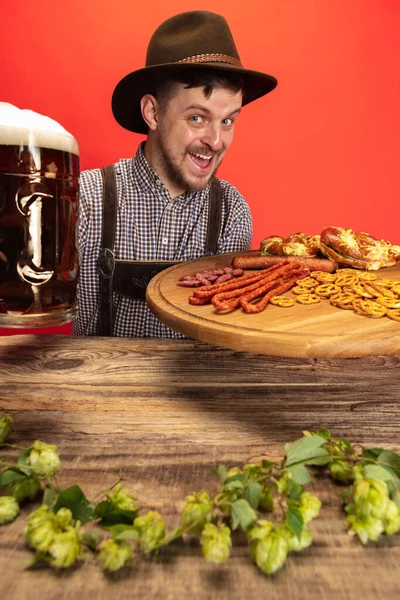 Счастливый улыбающийся мужчина, одетый в традиционный австрийский или баварский костюм, сидит за столом с праздничной едой и пивом, изолированным на красном фоне — стоковое фото