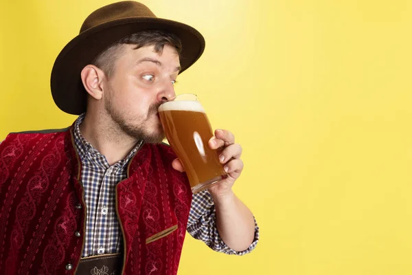 Close-up young man dressed in traditional Bavarian costume drinking cold foamed beer, lager, ale isolated over yellow background. National cuisine, holidays, traditions — Stock Photo, Image