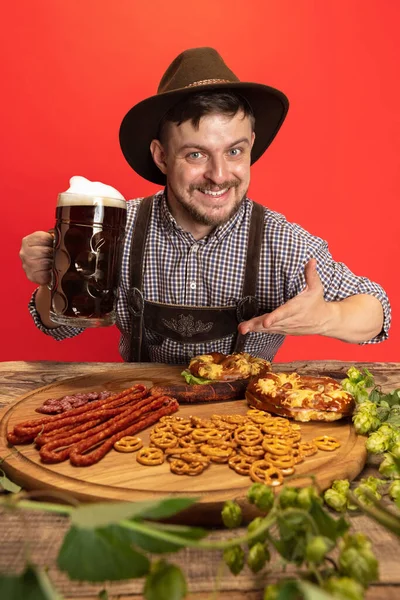 Feliz hombre sonriente vestido con traje tradicional austriaco o bávaro sentado a la mesa con comida festiva y cerveza aislada sobre fondo rojo —  Fotos de Stock