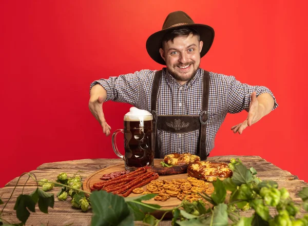 Glad leende man klädd i traditionella österrikiska eller bayerska kostym sitter vid bordet med festlig mat och öl isolerad över röd bakgrund — Stockfoto