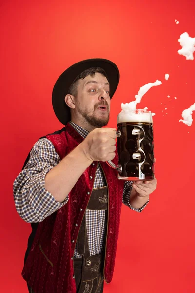 Happy smiling man dressed in traditional Austrian or Bavarian costume holding mug, glass of cold foamed dark black beer isolated over red background — Stock Photo, Image