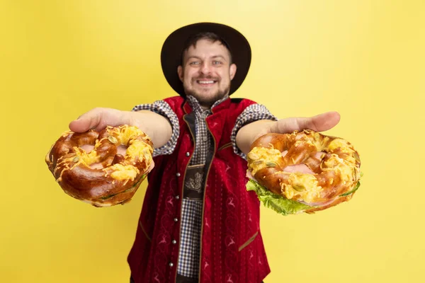 Щасливий усміхнений чоловік, одягнений в традиційний баварський костюм з юмоними кренделями, ізольований на жовтому тлі. Національна кухня, свята, традиції — стокове фото
