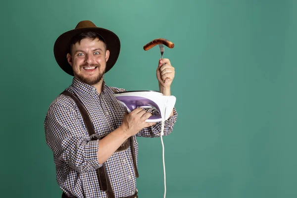 Comic portrait of young man in traditional Austrian or Bavarian costume frying a sausage on iron isolated over green background. Festival, humor concept — Stock Photo, Image