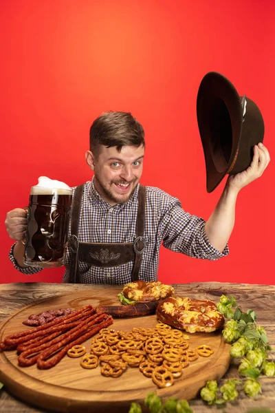 Gelukkig glimlachende man gekleed in traditionele Oostenrijkse of Beierse kostuum zitten aan tafel met feestelijk eten en bier geïsoleerd over rode achtergrond — Stockfoto