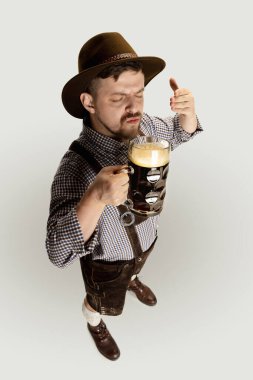 Sakallı adamın yüksek açılı görüntüsü, geleneksel Avusturya ya da Bavyera kostümlü garson elinde fincanla gri arka planda izole edilmiş koyu siyah bira bardağı.