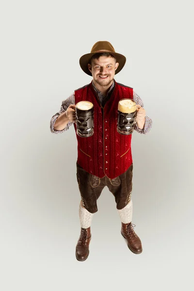 Feliz hombre sonriente vestido con traje tradicional austriaco o bávaro sosteniendo dos tazas, vasos de cerveza negra oscura aislados sobre fondo rojo. Volante —  Fotos de Stock