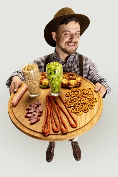 Feliz homem sorridente vestido com traje tradicional da Baviera segurando bandeja de madeira redonda com comida festiva e copos de cerveja cheios de quente selvagem e cevada — Fotografia de Stock