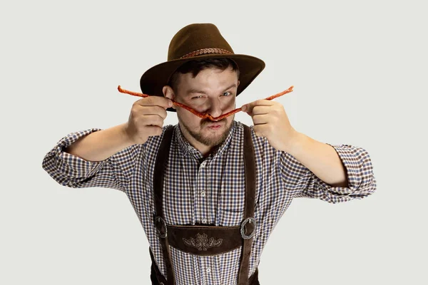 Słodki brodaty mężczyzna, kelner w tradycyjnym austriackim lub bawarskim kostiumie trzymający wędzone kiełbaski odizolowane na szarym tle. Zabawne meme emocje — Zdjęcie stockowe