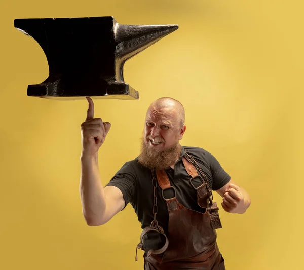 Comic portrait of muscular bearded bald man, blacksmith in leather apron or uniform isolated on yellow studio background. Concept of labor, retro professions, power, strength, humor — Stock Photo, Image