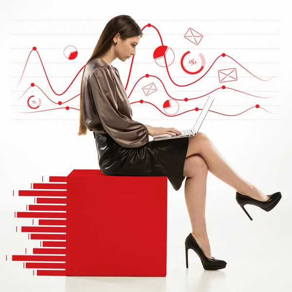 Mladá seriózní dívka, účetní, finanční analytička nebo úřednice v kancelářském obleku pomocí notebooku sedí na obrovském červeném čtverci, grafy izolované na světlém pozadí. — Stock fotografie