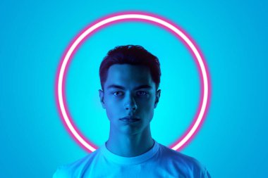 Sakin, kendinden emin. Genç adam portresi mavi stüdyo arka planında çok renkli neon ışıkla izole edilmiş geometrik ışıltılı şekil çemberiyle. İnsan duyguları kavramı, yüz ifadesi, reklam, moda.