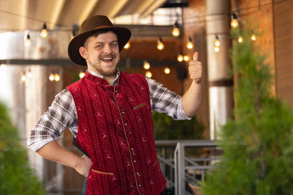 Happy smiling man, waiter in traditional Austrian or Bavarian costume holding mug, glass of dark beer and greeting customers at bar, cafe, pub. Oktoberfest — Stock Photo, Image