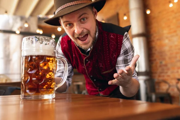 Happy smiling man, waiter in traditional Austrian or Bavarian costume holding mug, glass of dark beer and greeting customers at bar, cafe, pub. Oktoberfest — Stock Photo, Image