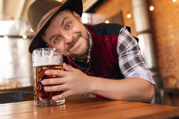 Happy smiling man, waiter in traditional Austrian or Bavarian costume holding mug, glass of dark beer and greeting customers at bar, cafe, pub. Oktoberfest — Stock Photo, Image