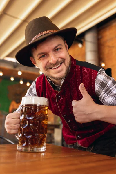 Happy smiling man, waiter in traditional Austrian or Bavarian costume holding mug, glass of dark beer and greeting customers at bar, cafe, pub. Oktoberfest — Stock Photo, Image