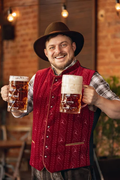 Feliz hombre sonriente, camarero con traje tradicional austriaco o bávaro sosteniendo taza, vaso de cerveza oscura y saludando a los clientes en el bar, cafetería, pub. Oktoberfest —  Fotos de Stock