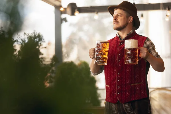 Happy smiling man, waiter in traditional Austrian or Bavarian costume holding mug, glass of dark beer and greeting customers at bar, cafe, pub. Oktoberfest — Stock Photo, Image