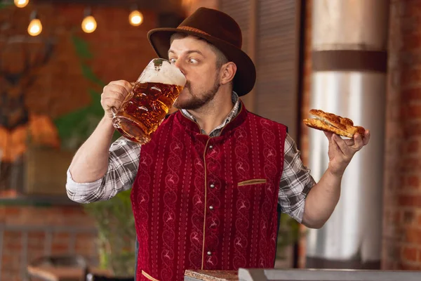 Portrait of happy bearded man in traditional Bavarian festive costume, hat and red vest tasting beer and snack during beer festival at bar, cafe, pub. Oktoberfest, october, autumn — Stock Photo, Image