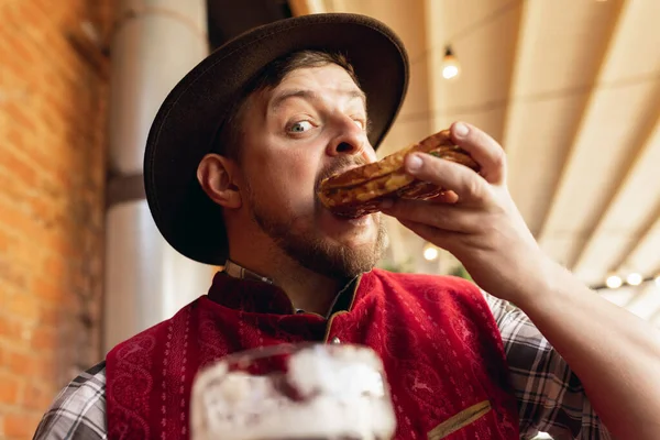 Portrait of happy bearded man in traditional Bavarian festive costume, hat and red vest tasting beer and snack during beer festival at bar, cafe, pub. Oktoberfest, october, autumn — Stock Photo, Image