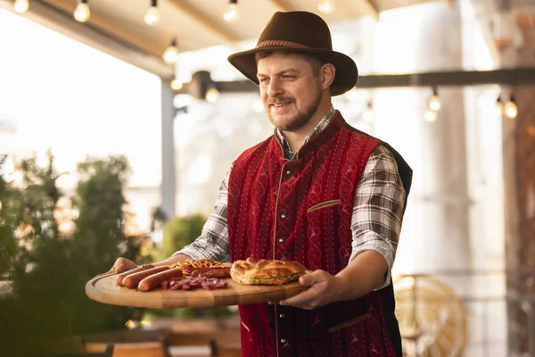 Happy smiling man, waiter dressed in traditional Bavarian costume holding round wooden tray with festive food, pretzel and sausages. Oktoberfest, holidays, autumn vibe
