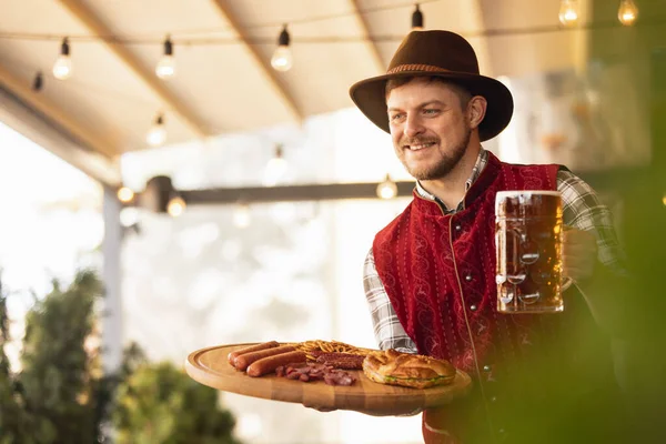 Feliz hombre sonriente, camarero con traje tradicional austriaco o bávaro sosteniendo taza, vaso de cerveza oscura y saludando a los clientes en el bar, cafetería, pub. Oktoberfest — Foto de Stock