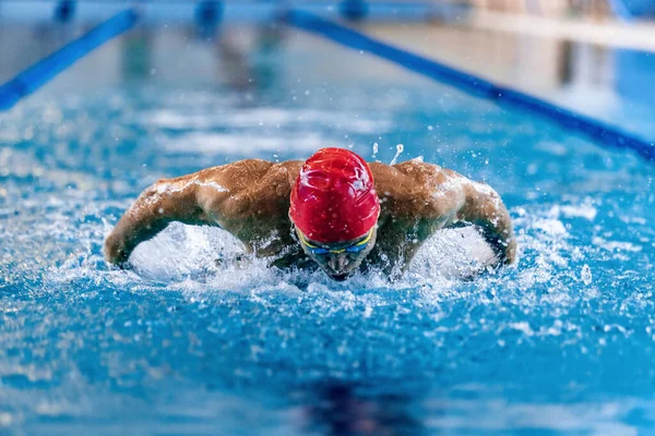 Vista frontal do nadador masculino profissional em boné vermelho e óculos em movimento e ação durante o treinamento na piscina, dentro de casa. Estilo de vida saudável, poder, energia, conceito de movimento desportivo — Fotografia de Stock