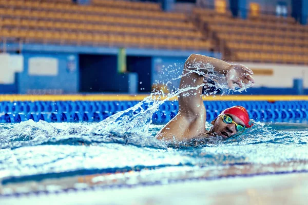 Nadador masculino profissional em touca de natação e óculos em movimento e ação durante o treinamento na piscina, dentro de casa. Estilo de vida saudável, poder, energia, conceito de movimento desportivo — Fotografia de Stock