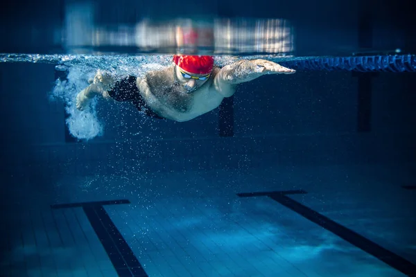 Nadador masculino profesional en gorra de natación y gafas en movimiento y acción durante el entrenamiento en la piscina, en interiores. Estilo de vida saludable, poder, energía, concepto de movimiento deportivo —  Fotos de Stock