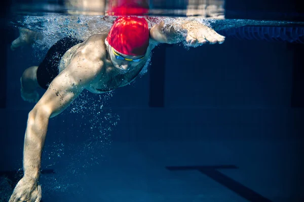 Underwater view of professional male swimmer in red cap and goggles in motion and action during training at pool, indoors. Healthy lifestyle, power, energy, sports movement concept — Stock Photo, Image