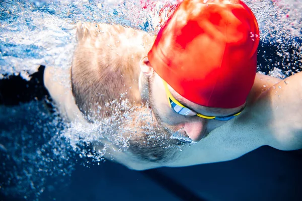 Close-up. Vista subaquática do nadador masculino profissional em boné vermelho e óculos em movimento e ação durante o treinamento na piscina, dentro de casa. Estilo de vida saudável, poder, energia, conceito de movimento desportivo — Fotografia de Stock