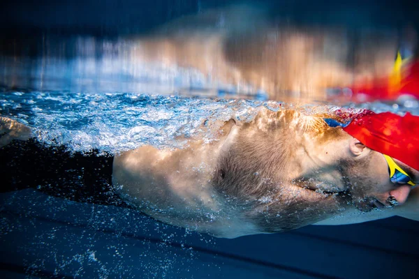 Крупный план. Подводный вид профессионального пловца в красной кепке и очки в движении и действии во время тренировок в бассейне, в помещении. — стоковое фото