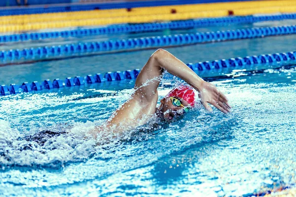 Nadador masculino profissional em touca de natação e óculos em movimento e ação durante o treinamento na piscina, dentro de casa. Estilo de vida saudável, poder, energia, conceito de movimento desportivo — Fotografia de Stock