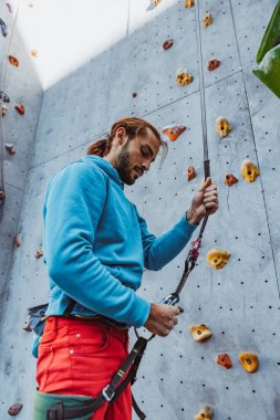 Tırmanma hazırlığı. Genç adam profesyonel kaya tırmanışçısı güneşli bir günde, açık havada eğitim merkezine tırmanmadan önce spor malzemelerini kontrol ediyor. Sağlıklı aktif yaşam tarzı kavramı, meydan okuma