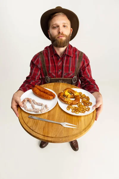 Vista superior del joven barbudo, camarero en traje bávaro tradicional sosteniendo bandeja de madera redonda con comida festiva, aperitivos, salchichas y pretzels. Oktoberfest — Foto de Stock