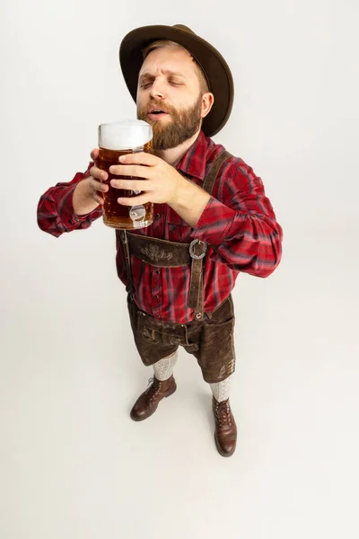 Half-length portrait of bearded man in hat and traditional Bavarian costume holding huge mug, glass of light frothy beer isolated over white background. Flyer — Stock Photo, Image