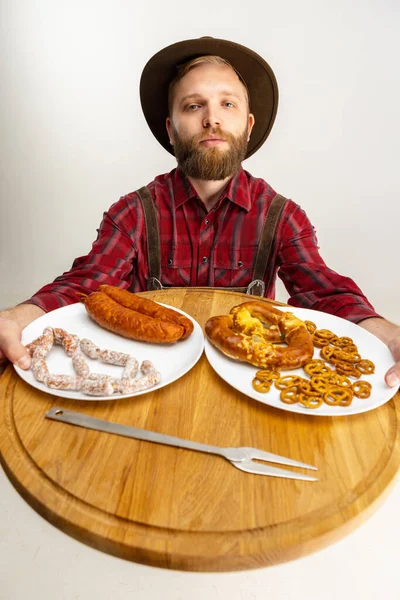 Top view of young bearded man, waiter in traditional Bavarian costume holding round wooden tray with festive food, snacks, sausages and pretzels. Oktoberfest