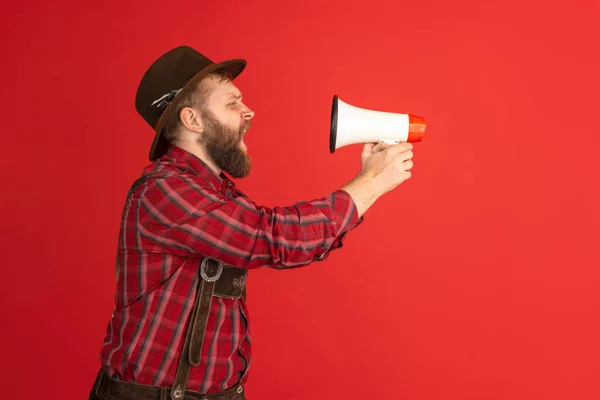 Ritratto comico di uomo barbuto, cameriere in cappello e costume tradizionale bavarese che grida al megafono isolato su sfondo rosso da studio. Oktoberfest, fest, concetto di vacanza — Foto Stock